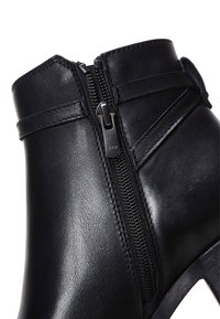 Bottine en cuir noir avec fermeture éclair latérale apparente et détail de sangle décorative sur une surface lisse et texturée.