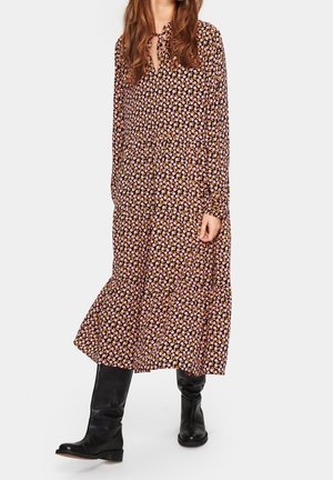 Nainen pukeutuneena pitkähihainen, nilkkoihin ulottuva kukallinen mekko, jossa on pieniä vaaleanpunaisia ja oransseja kukkia, yhdistettynä mustiin nahkaisiin polvipitkiin saappaisiin.