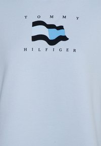 Sudadera azul claro con un gráfico de una bandera ondeando en negro, blanco y azul claro, con el texto "TOMMY HILFIGER" debajo. Textura suave.