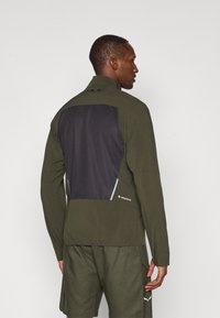 Πράσινο αθλητικό μπουφάν με μαύρη διχτυωτή πίσω πάνελ, ψηλό γιακά και μακριά μανίκια, κατασκευασμένο από ελαφρύ, ευέλικτο υλικό.