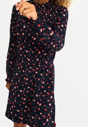 Barn iført en marineblå kjole med opsmøgede manchetter og krave, med et rødt og pink blomsterprint på hvid baggrund.