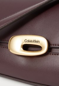 Sac à main Calvin Klein en cuir bourgogne profond avec un fermoir doré à finition brillante et un évidement ovale, ainsi qu'un logo de marque embossé. Texture lisse.