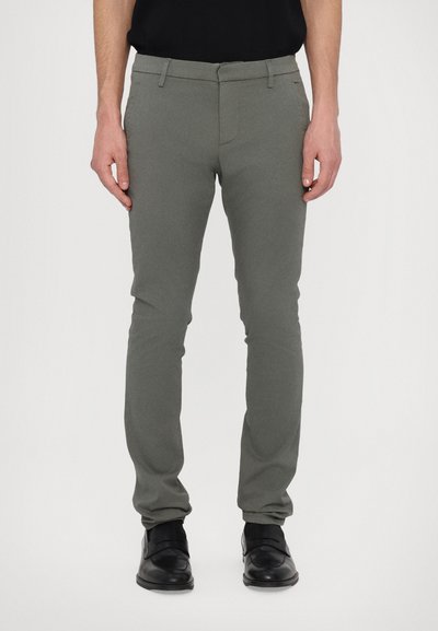Pantalon gris slim fit en tissu doux avec une texture lisse ; possède des passants de ceinture et une fermeture éclair. Porté avec des chaussures noires.