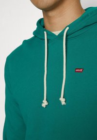 Homme portant un sweat à capuche Levi's bleu sarcelle avec des cordons blancs et le logo Levi's visible sur la poitrine, vue en gros plan du cou à la poitrine.