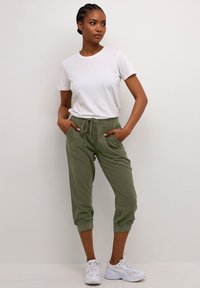 Maglietta bianca a maniche corte abbinata a pantaloni della tuta verde oliva tagliati sopra la caviglia, con coulisse regolabile e tasche laterali, indossata con sneakers bianche.