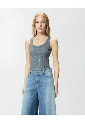 Giovane donna con lunghi capelli biondi, indossa un top grigio senza maniche con borchie e jeans larghi azzurri, in piedi davanti a uno sfondo bianco uniforme.