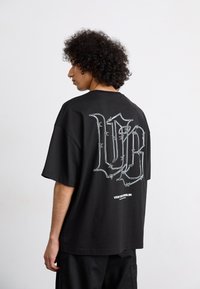 Sort oversized t-shirt med et stort hvidt grafisk design med sammenflettede bogstaver på ryggen. Blødt stof, afslappet pasform, korte ærmer.