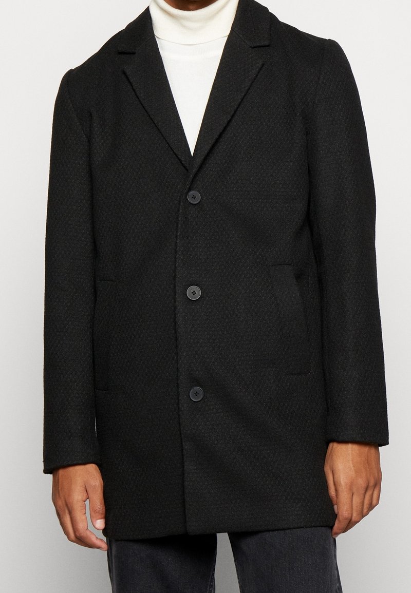 Homme portant un manteau noir texturé boutonné par-dessus un pull à col roulé blanc, debout les bras détendus le long du corps.