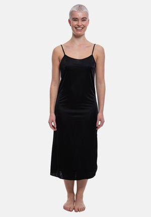 Lächende Person mit kurzen blonden Haaren, steht barfuß und trägt ein ärmelloses schwarzes Midikleid mit dünnen Trägern vor einem schlichten hellen Hintergrund.