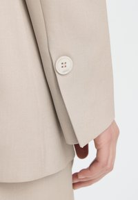 Beige tailored suit jacket sleeve with a smooth texture, featuring a single round, four-hole button near the cuff.