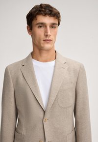 Blazer in tessuto beige con texture a spina di pesce, taschino singolo e chiusura a due bottoni, indossato sopra una camicia bianca.