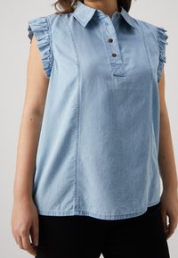 Top in denim azzurro con colletto, patta dei bottoni e maniche con volant. Presenta cuciture verticali e una vestibilità comoda.