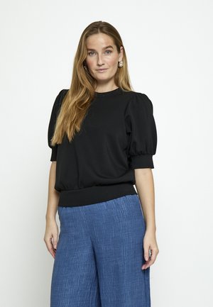 Kobieta z długimi blond włosami, ubrana w czarną bluzkę z bufiastymi rękawami i luźne niebieskie spodnie, stojąca na tle jednolitego jasnego tła.