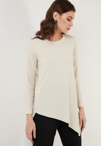 Maglia a maniche lunghe color crema con finitura texturizzata, caratterizzata da un orlo asimmetrico e spacchi laterali. Abbinata a pantaloni neri, con decorazioni minime.