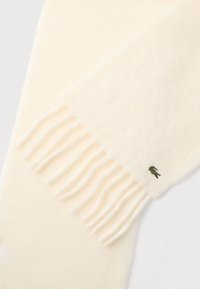 Bufanda de lana color crema con extremos deshilachados. Presenta un pequeño logo bordado en verde. Textura suave con diseño minimalista.