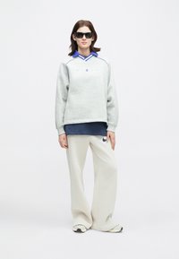 Γυναίκα που φορά μαύρα γυαλιά ηλίου, ανοιχτό γκρι φούτερ πάνω από ναυτικό μπλε πουκάμισο με γιακά, κρεμ παντελόνια σε φαρδύ σχέδιο και λευκά αθλητικά παπούτσια, που στέκει σε άσπρο φόντο.