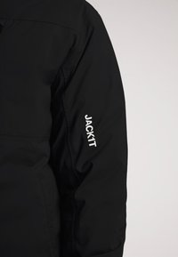 Giacca nera con una superficie liscia e chiusura con zip. Presenta il logo bianco "JACKIT" sulla manica sinistra. Materiale leggero.