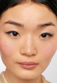 Närbild av ett ansikte med slät, lätt fräknig hud, rosa rouge och djärv blå eyeliner. Neutrala läppar och mörkt hår, med ett fokuserat uttryck.