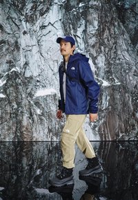 Veste imperméable navy, pantalon cargo beige et bottes de randonnée noires. Les caractéristiques incluent un devant zippé, une capuche ajustable et plusieurs poches.