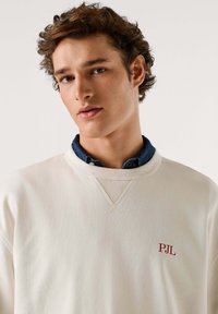 Felpa color crema con collo e polsini a costine, con ricamo rosso "PJL" sul petto, indossata sopra una camicia di jeans blu.