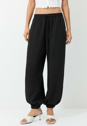 Femme portant un pantalon jogger noir avec taille et poignets élastiques, associé à des sandales blanches à talons et un top court blanc.