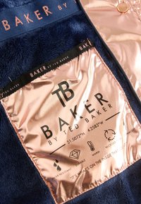 Tissu bleu marine avec étiquette brillante en or rose indiquant Baker by Ted Baker, avec des icônes de matériaux résistants à l'eau, durables, chauds et recyclés.