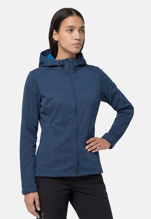 Donna che indossa una giacca softshell blu con cappuccio, cerniera frontale e tasche laterali, in piedi con una mano sui fianchi.