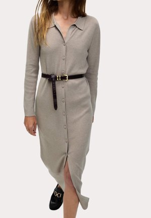 Kvinde iført en beige midi-kjole med knapper og krave, et sort bælte og sorte loafers med gyldne detaljer, går fremad.