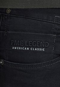 Primo piano di una tasca di jeans neri in denim con testo in rilievo "PME Legend" e testo stampato "American Classic".