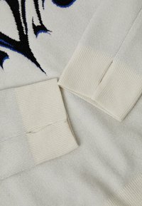 Λευκό πλεκτό ύφασμα με ριμπ μανσέτες. Το σχέδιο περιλαμβάνει ένα μαύρο και μπλε μοτίβο, τονίζοντας την υφή και τη λεπτομέρεια στο υλικό.