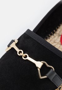 Chaussure plate en daim noir avec un accent en chaîne dorée et des brides en cuir noir. La semelle présente une texture tissée naturelle.