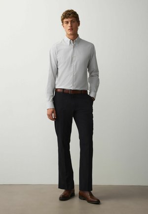 Fiatal férfi áll, világosszürke gombos inget, fekete nadrágot, barna övet és barna bőr cipőt visel, egyszínű fehér háttér előtt.