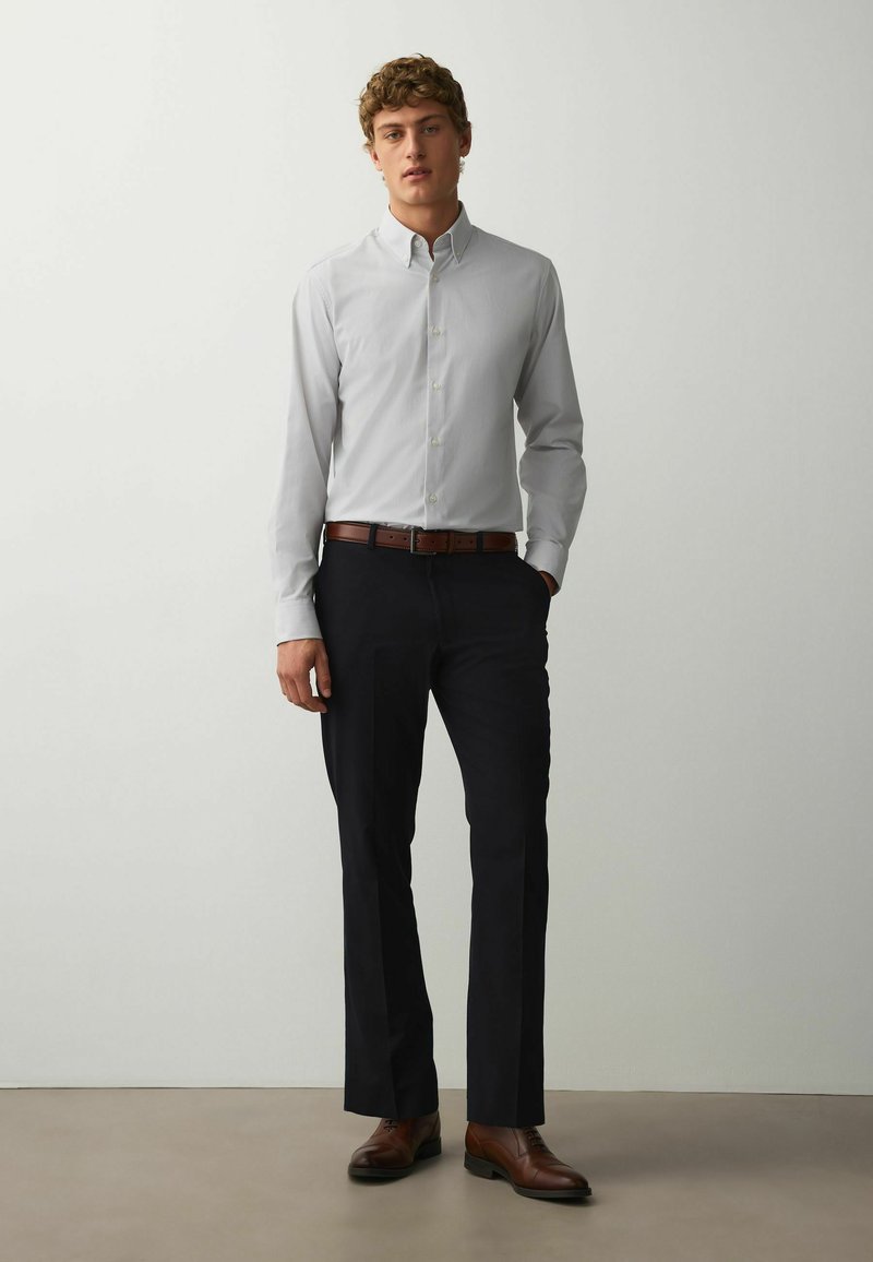 Jeune homme debout portant une chemise gris clair à boutons, un pantalon noir, une ceinture marron et des chaussures en cuir marron sur un fond blanc uni.
