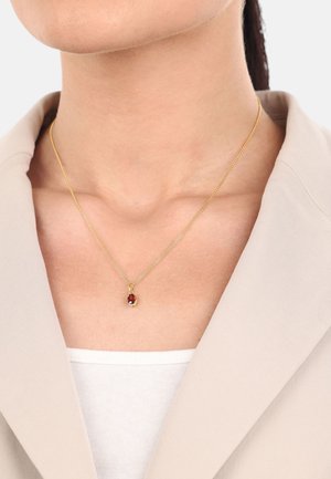 Collar de cadena de oro con un colgante en forma de lágrima roja con un acento negro, mostrado en una persona que lleva puesto un blazer beige y una parte superior blanca.