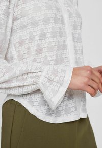 Witte kanten blouse met een gemêleerd bloemenpatroon, knoopsluiting aan de voorkant en gerimpelde manchetten; gecombineerd met olijfgroene broeken voor een mooi contrast.