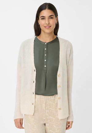 Lichtbeige cardigan van zacht materiaal, met knopenvoorzijde met houten knopen en een open breistekstuur, gedragen over een donkergroene blouse.