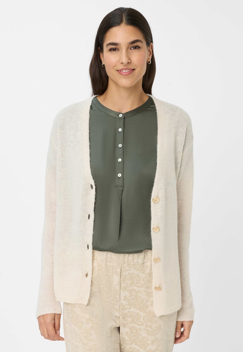 Helle beige Strickjacke aus weichem Material, mit einer Knopfleiste aus Holzknöpfen und offener Maschenstruktur, getragen über einer dunkelgrünen Bluse.