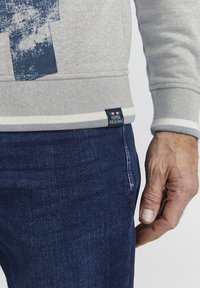 Grauer Pullover mit blauem Grafikdesign, gerippten Bündchen und Saum mit weißen Streifen; kombiniert mit dunklen Jeans. Markenlabel sichtbar.