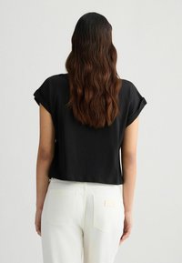 Top noir court avec des manches courtes repliées, accompagné d'un pantalon blanc taille haute. Texture lisse, design minimaliste. Vue de dos.