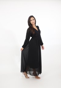 Kobieta w długiej czarnej sukni z długimi rękawami trzyma rąbek sukni, nosząc przezroczyste sandały na wysokim obcasie, stojąca na białym tle.