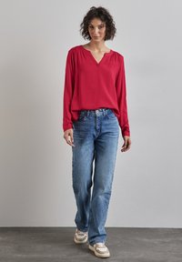 Blusa rossa a maniche lunghe con scollatura a V, spalle arricciate, abbinata a jeans in denim blu larghi e scarpe da ginnastica beige. Texture del tessuto liscia.