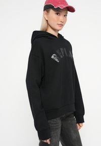Černá mikina s cropped designem, se lesklým logem "REPLAY". Model má na hlavě růžovou čepici, která zvýrazňuje ležérní vzhled outfitu.