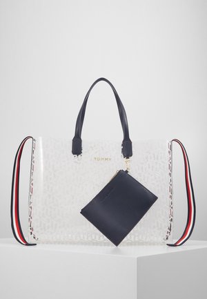 Sac fourre-tout transparent avec poignées bleu marine, bandoulière amovible rayée rouge-blanc-bleu, logo doré « TOMMY » et pochette bleu marine attachée.