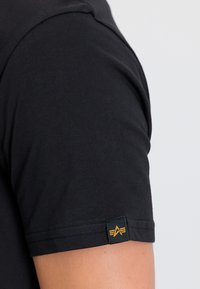 Must mustru, millel on lühikesed varrukad, pehme tekstuur ja väike roheline silt, millel on kollane logo detail varruka peal.