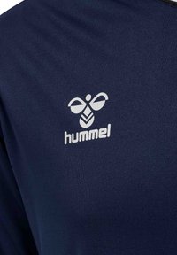 Weißes Hummel-Markenlogo mit Bienen-Symbol auf dunkelblauem Stoff, wahrscheinlich Sport- oder Activewear.