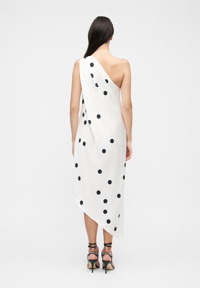 Γυναίκα με μακριά σκούρα μαλλιά που φοράει λευκό φόρεμα με έναν ώμο και μαύρα πουά, και μαύρα ψηλοτάκουνα σανδάλια, με την πλάτη στραμμένη.