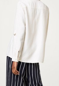 Chemise en lin blanche à coupe décontractée, avec des poignets à boutons et une couture de yoke au dos. Associée à un pantalon à rayures bleu marine.
