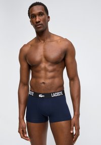 Αθλητικός άντρας με γυμνό κορμό που φοράει μπλε ναυτικό μποξεράκια Lacoste, σε απλό φωτεινό φόντο.