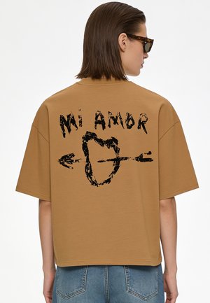 Person mit kurzen braunen Haaren, die Sonnenbrillen trägt und ein braunes Hemd mit "Mi Amor" und einem Herz, das von einem Pfeil durchbohrt ist, auf dem Rücken.