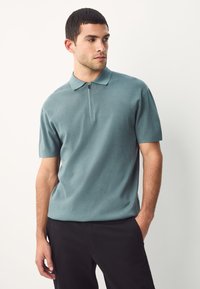 Kurzärmeliges Poloshirt in gedämpftem Türkis mit Reißverschlusskragen, strukturierter Stoff und lockerer Passform, kombiniert mit dunkler Hose.